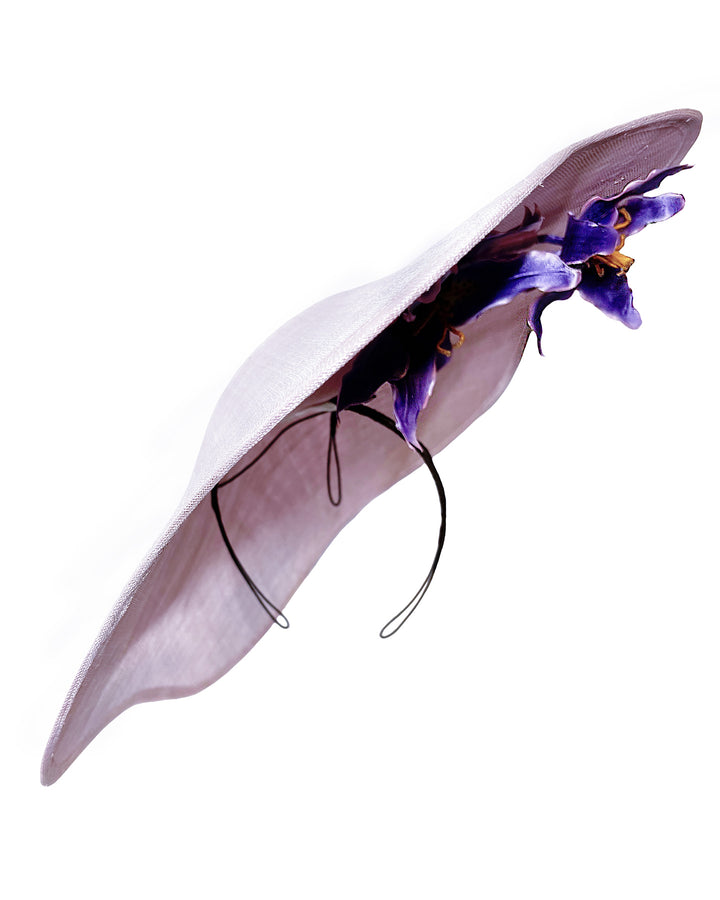 Lilac sinamay wide-brim fascinator with a softly curved sculptural silhouette, featuring striking purple floral accents nestled beneath the brim for a bold yet refined finish. Designed and made by Merve Bayindir Millinery.
