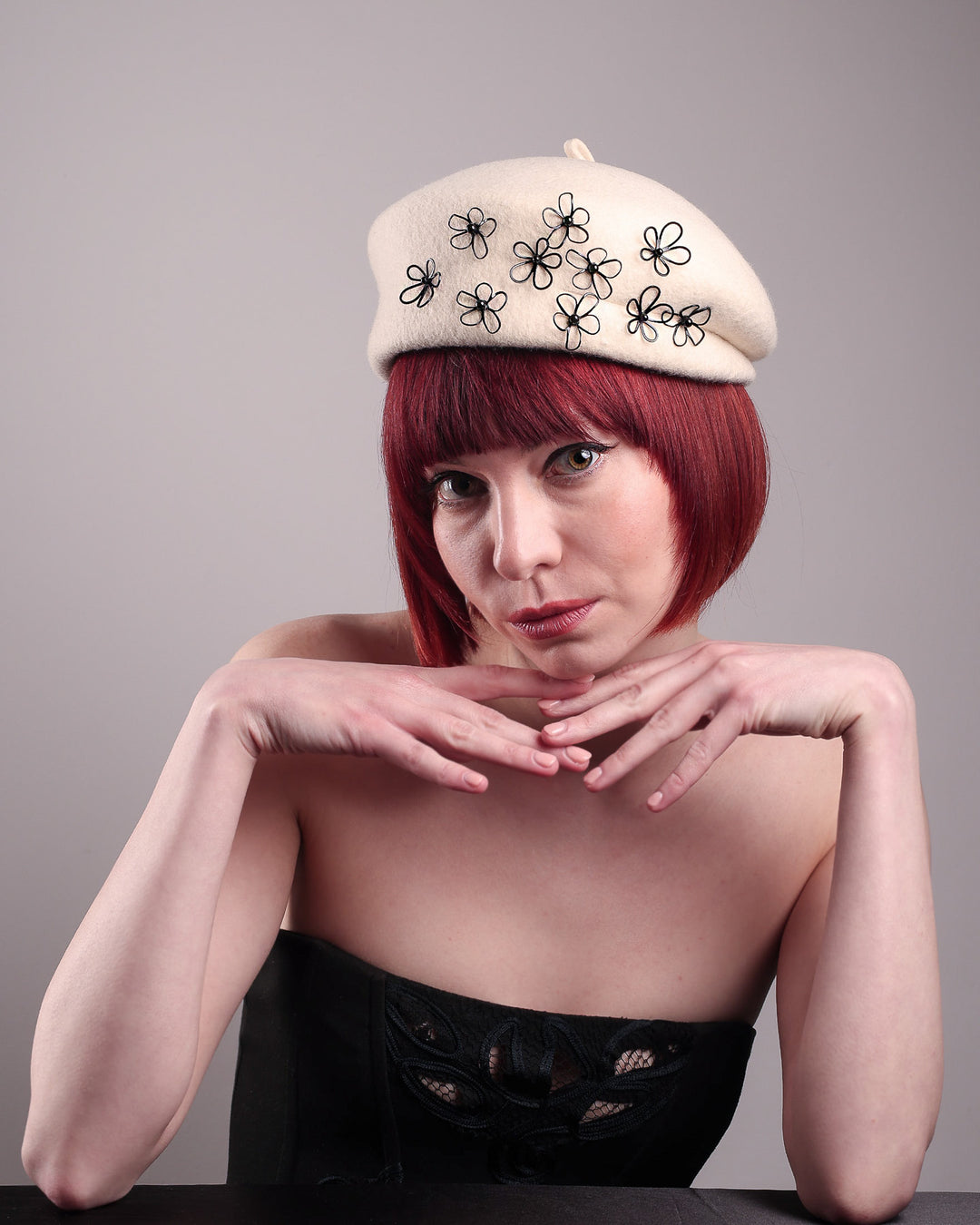The model, showcasing the front of the Vitra Beret, crafted from soft white felt, features a scattering of hand-applied black wire flowers on the front to left side that seem to dance across the crown like doodles. Designed by Merve Bayindir.