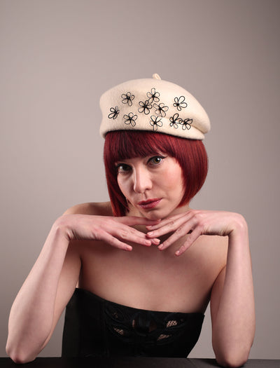 The model, showcasing the front of the Vitra Beret, crafted from soft white felt, features a scattering of hand-applied black wire flowers on the front to left side that seem to dance across the crown like doodles. Designed by Merve Bayindir.