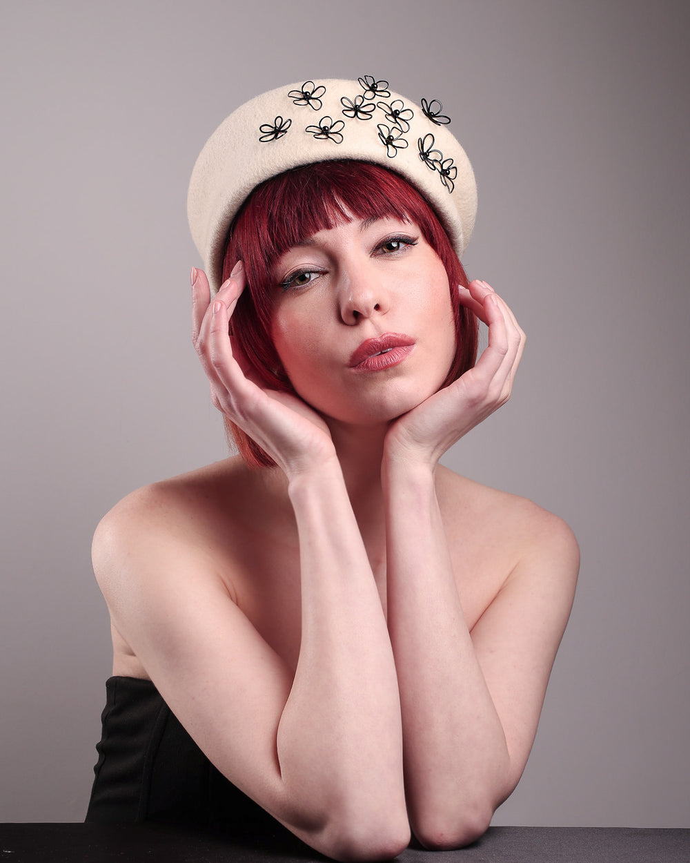 The model, showcasing the front from the below angle of the Vitra Beret, crafted from soft white felt, features a scattering of hand-applied black wire flowers on the front to left side that seem to dance across the crown like doodles. Designed by Merve Bayindir.
