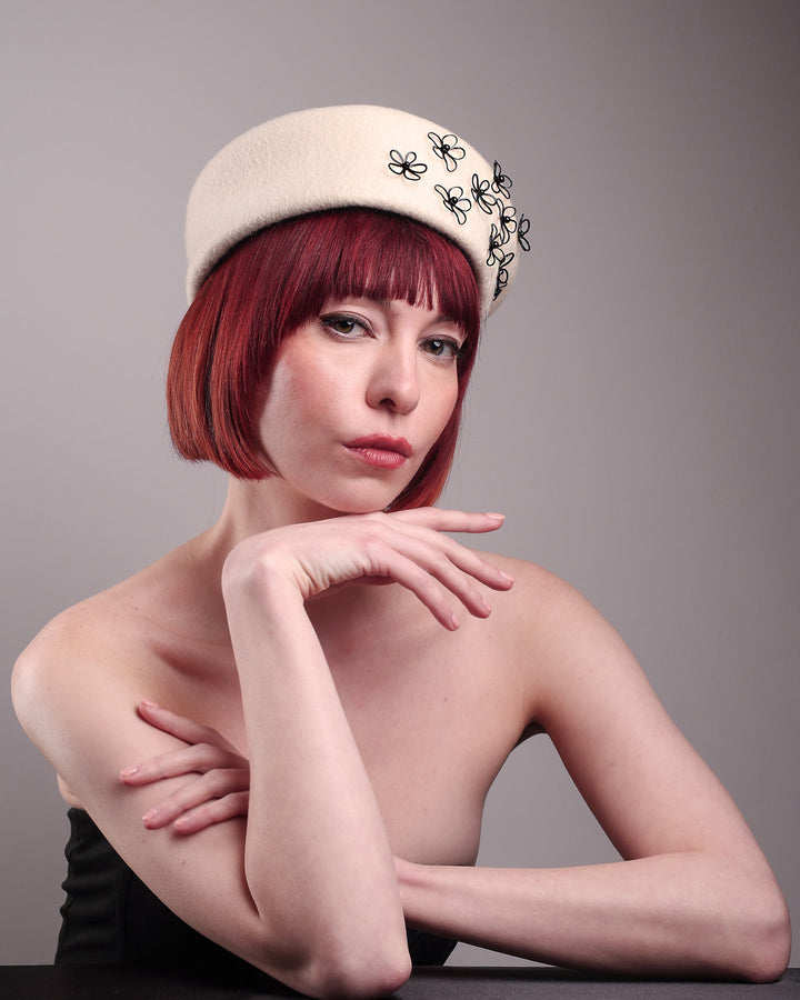 The model, showcasing the front right side of the Vitra Beret, crafted from soft white felt, features a scattering of hand-applied black wire flowers on the front to left side that seem to dance across the crown like doodles. Designed by Merve Bayindir.