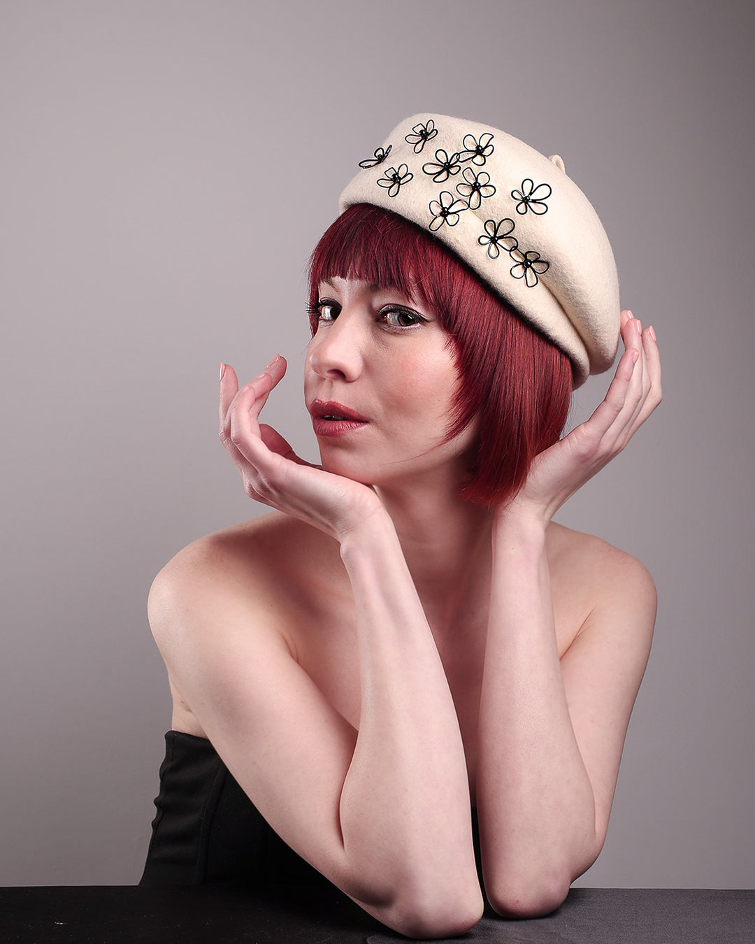 The model, showcasing the front left side of the Vitra Beret, crafted from soft white felt, features a scattering of hand-applied black wire flowers on the front to left side that seem to dance across the crown like doodles. Designed by Merve Bayindir.