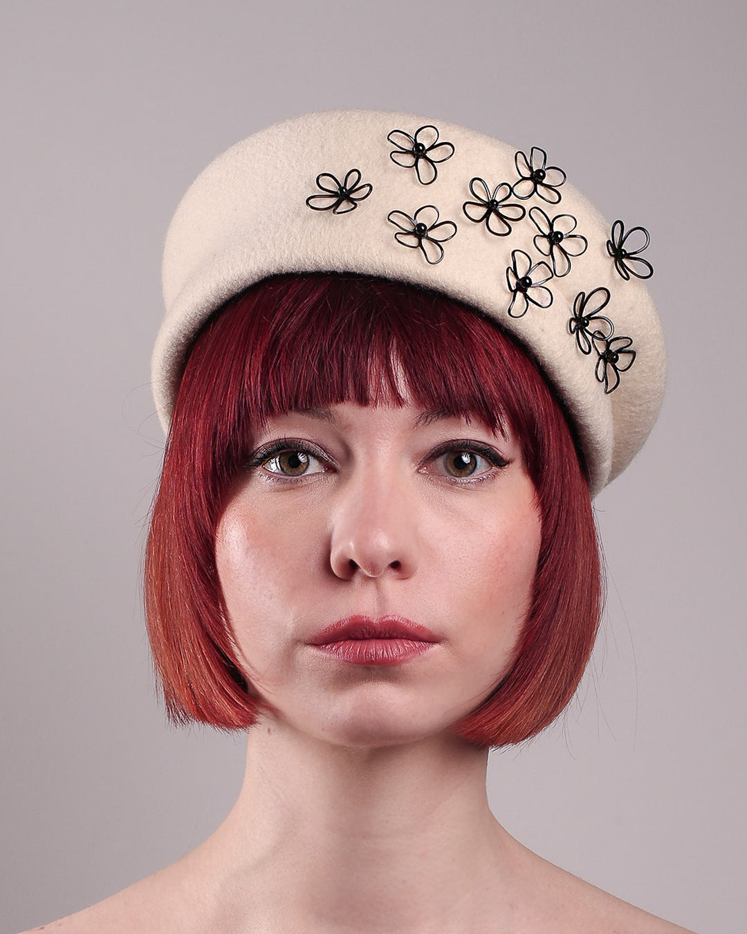 The model, showcasing the front close up of the Vitra Beret, crafted from soft white felt, features a scattering of hand-applied black wire flowers on the front to left side that seem to dance across the crown like doodles. Designed by Merve Bayindir.