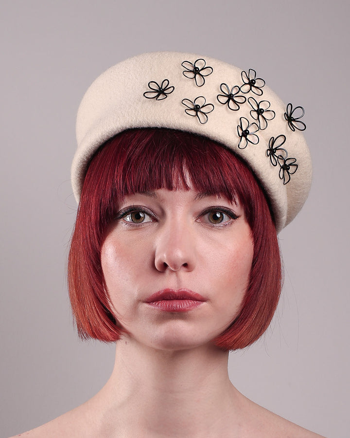 The model, showcasing the front close up of the Vitra Beret, crafted from soft white felt, features a scattering of hand-applied black wire flowers on the front to left side that seem to dance across the crown like doodles. Designed by Merve Bayindir.
