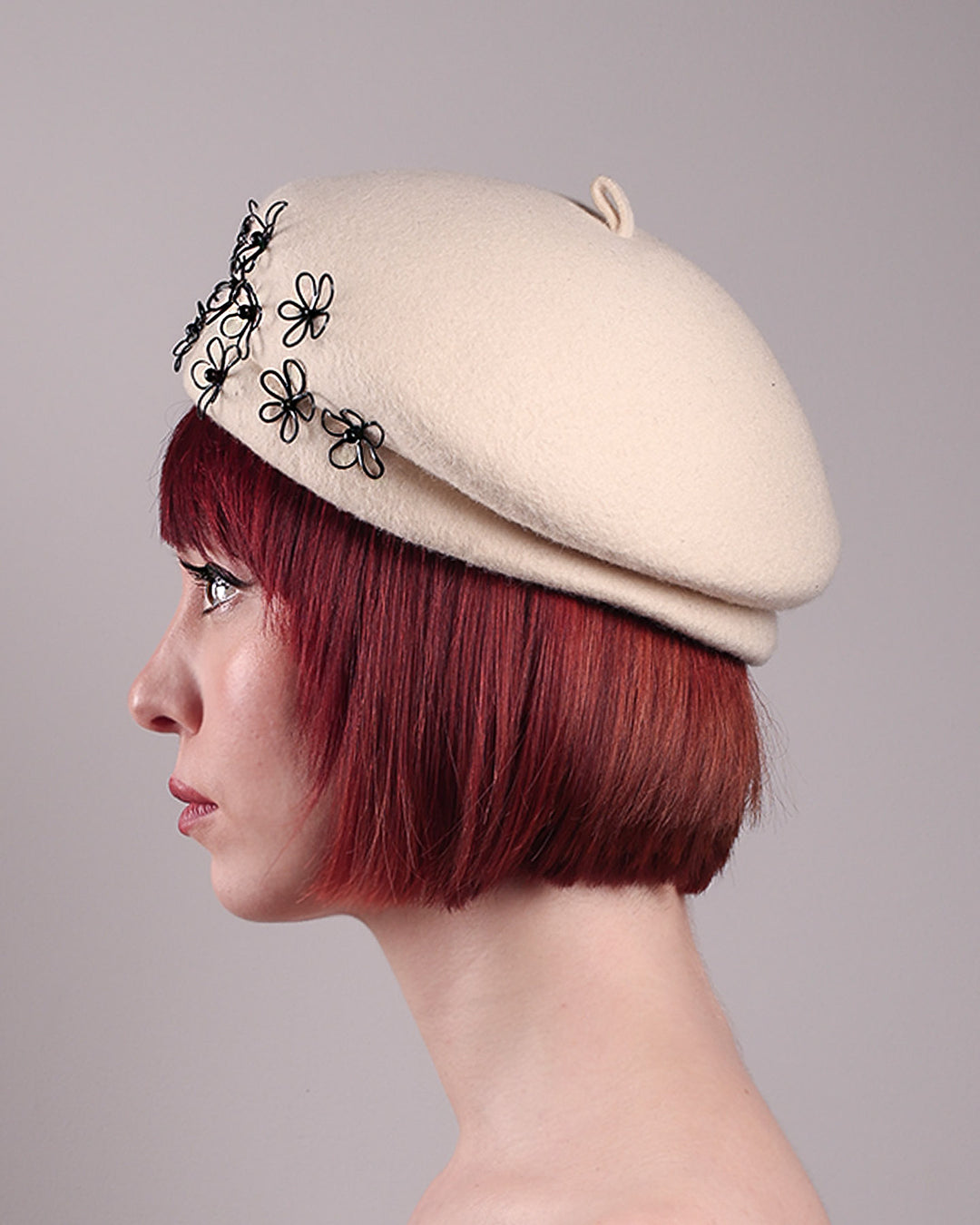The model, showcasing the left side close up of the Vitra Beret, crafted from soft white felt, features a scattering of hand-applied black wire flowers on the front to left side that seem to dance across the crown like doodles. Designed by Merve Bayindir.