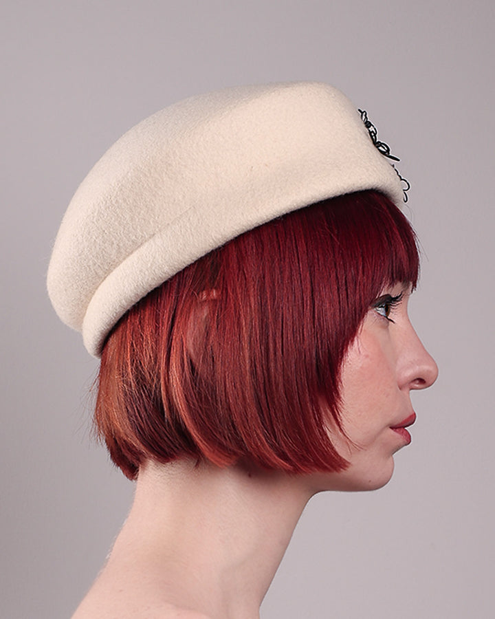 The model, showcasing the right side close up of the Vitra Beret, crafted from soft white felt, features a scattering of hand-applied black wire flowers on the front to left side that seem to dance across the crown like doodles. Designed by Merve Bayindir.