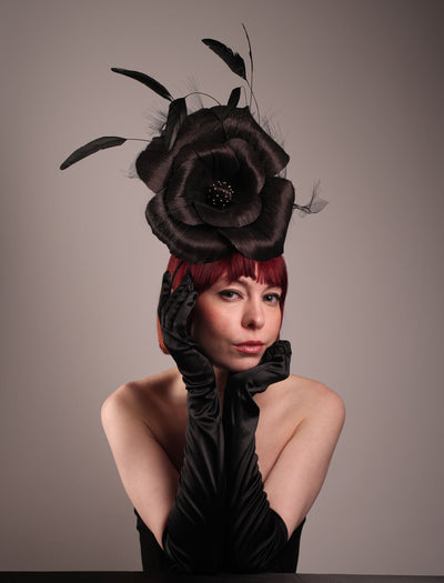 Model is wearing a large black silk abaca flower fascinator by Merve Bayindir. The black flower centre is embellished with gold beads. Back of the silk abaca black flower is decorated with long trimmed rooster feathers and crinoline cloud. The fascinator is fitted with a sleek headband.
