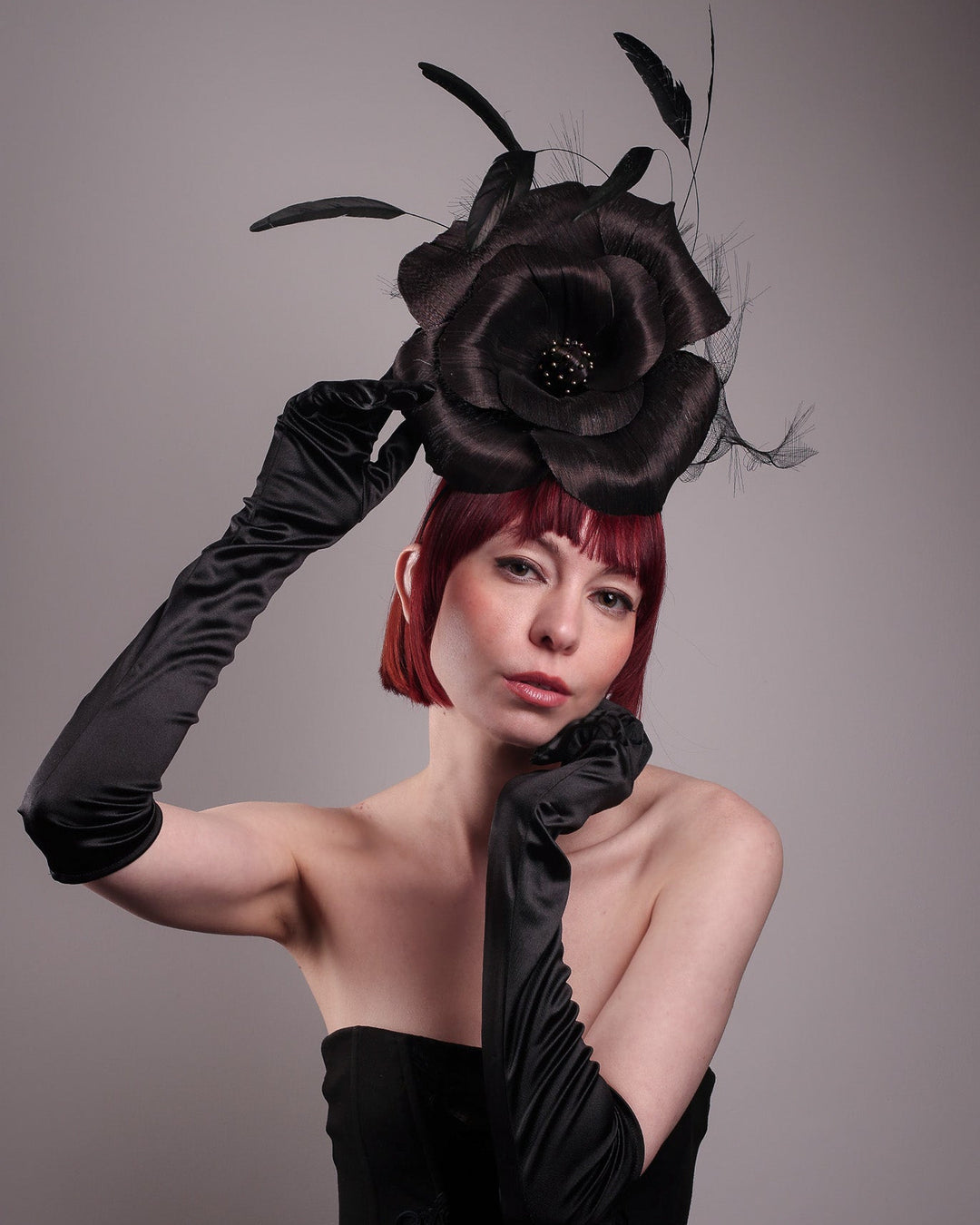 Model showcasing the front of a large black silk abaca flower fascinator by Merve Bayindir. The centre of the black flower is embellished with gold beads. Back of the silk abaca black flower is decorated with long trimmed rooster feathers and crinoline cloud. The fascinator is fitted with a sleek headband.
