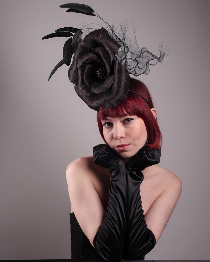 Model is showcasing the left frint side of  a large black silk abaca flower fascinator by Merve Bayindir. The centre of the black flower is embellished with gold beads. Back of the silk abaca black flower is decorated with long trimmed rooster feathers and crinoline cloud. The fascinator is fitted with a sleek headband.