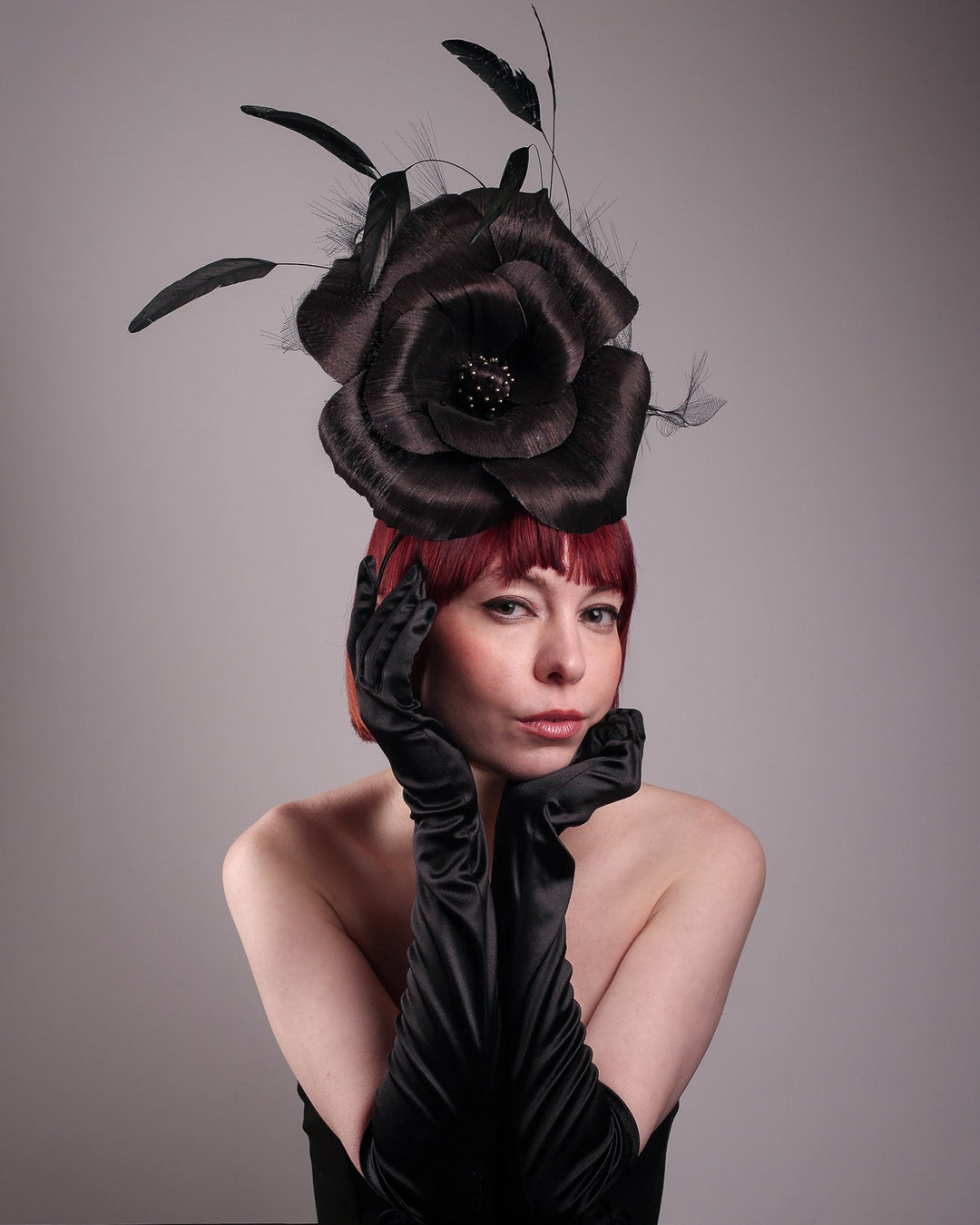 Model is wearing a large black silk abaca flower fascinator by Merve Bayindir. The black flower centre is embellished with gold beads. Back of the silk abaca black flower is decorated with long trimmed rooster feathers and crinoline cloud. The fascinator is fitted with a sleek headband.
