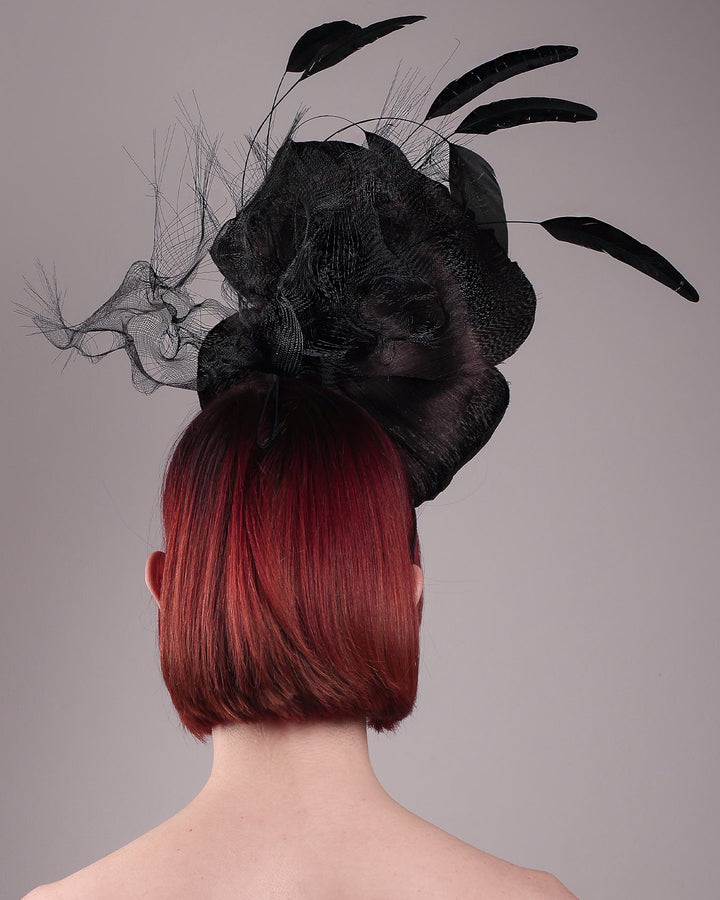 Product picture with model. Model showcasing back view of a large black silk abaca flower fascinator by Merve Bayindir. The centre of the black flower is embellished with gold beads. Back of the silk abaca black flower is decorated with long trimmed rooster feathers and crinoline cloud. The fascinator is fitted with a sleek headband.