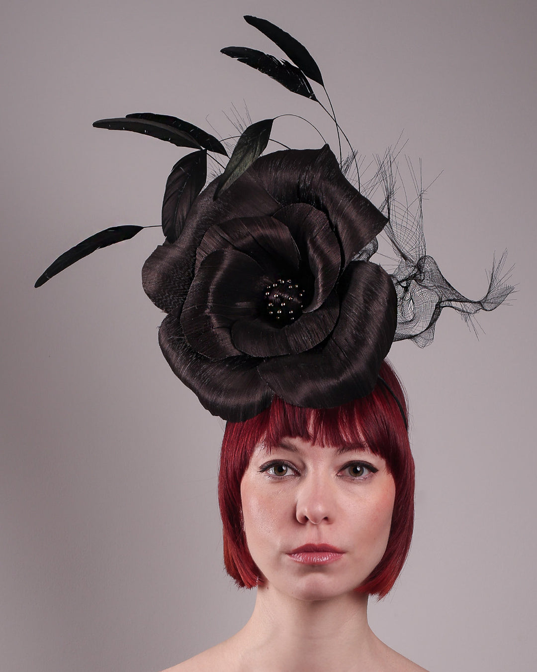 Product picture with model. Model showcasing front view of a large black silk abaca flower fascinator by Merve Bayindir. The centre of the black flower is embellished with gold beads. Back of the silk abaca black flower is decorated with long trimmed rooster feathers and crinoline cloud. The fascinator is fitted with a sleek headband.