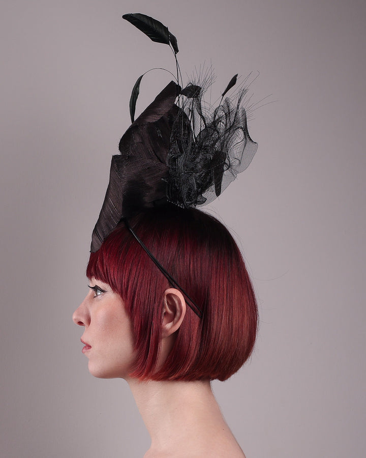 Product picture with model. Model showcasing left view of a large black silk abaca flower fascinator by Merve Bayindir. The centre of the black flower is embellished with gold beads. Back of the silk abaca black flower is decorated with long trimmed rooster feathers and crinoline cloud. The fascinator is fitted with a sleek headband.