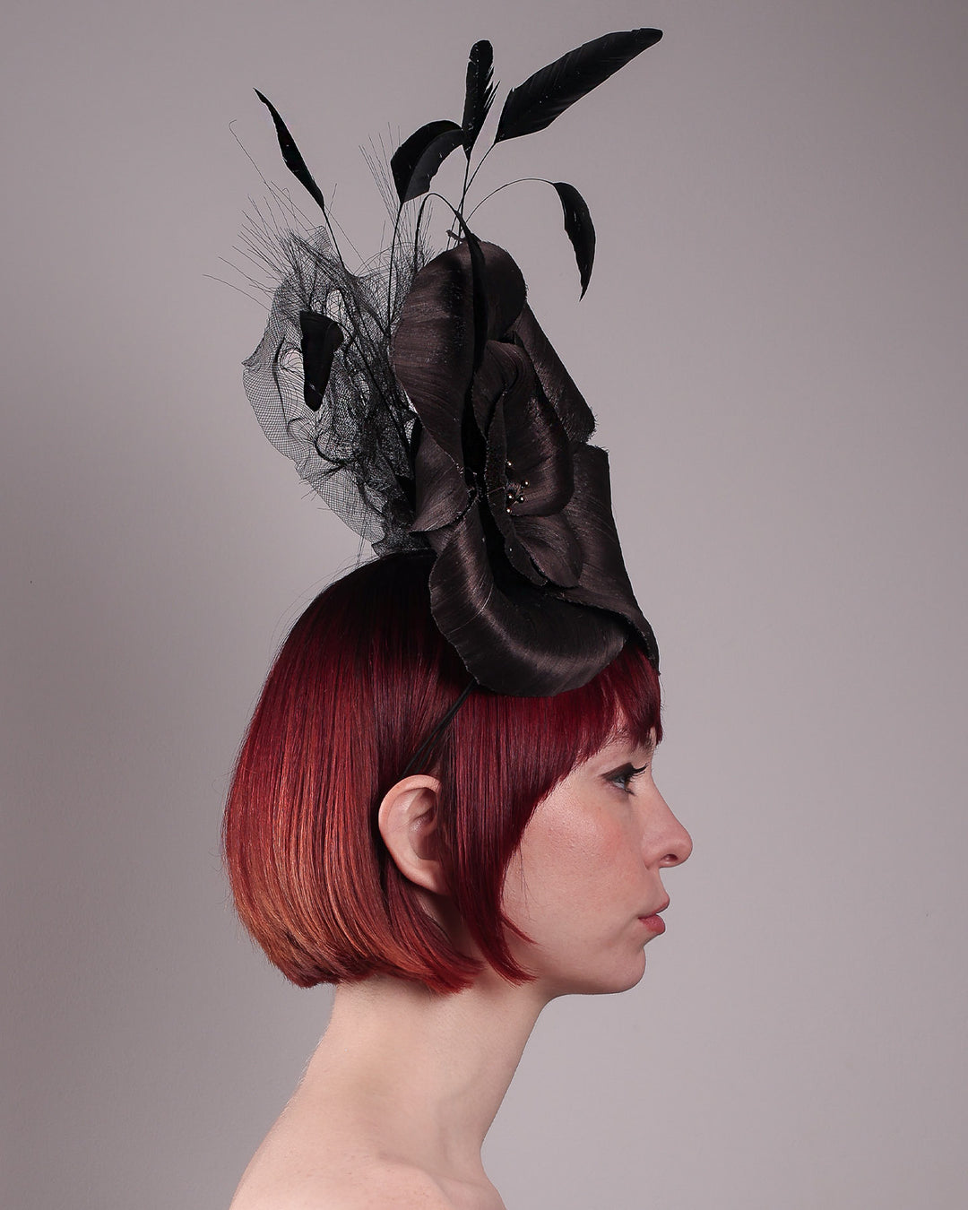 Product picture with model. Model showcasing right view of a large black silk abaca flower fascinator by Merve Bayindir. The centre of the black flower is embellished with gold beads. Back of the silk abaca black flower is decorated with long trimmed rooster feathers and crinoline cloud. The fascinator is fitted with a sleek headband.