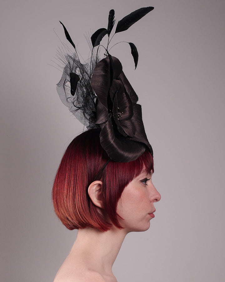 Product picture with model. Model showcasing right view of a large black silk abaca flower fascinator by Merve Bayindir. The centre of the black flower is embellished with gold beads. Back of the silk abaca black flower is decorated with long trimmed rooster feathers and crinoline cloud. The fascinator is fitted with a sleek headband.