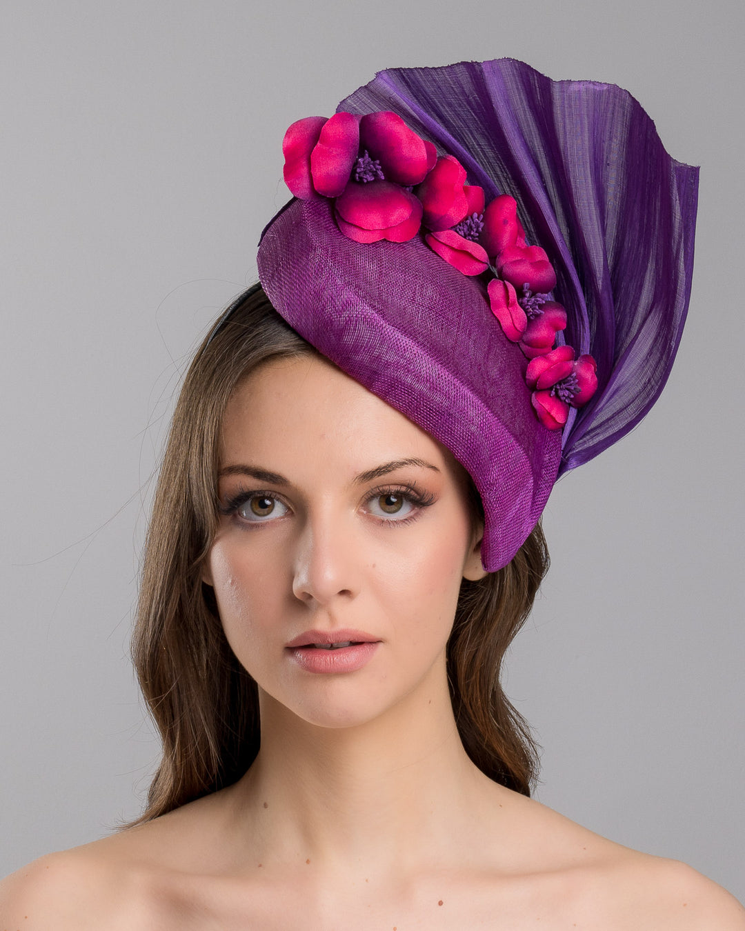 The model is showcasing the front side close up of the West is a purple side percher style fascinator curving to the left of the head framing the right side of the face. Decorated with a variety of pink and purple shaded cherry flowers and completed with a purple pleaded fan in the back. Designed and made by Merve Bayindir Millinery.
