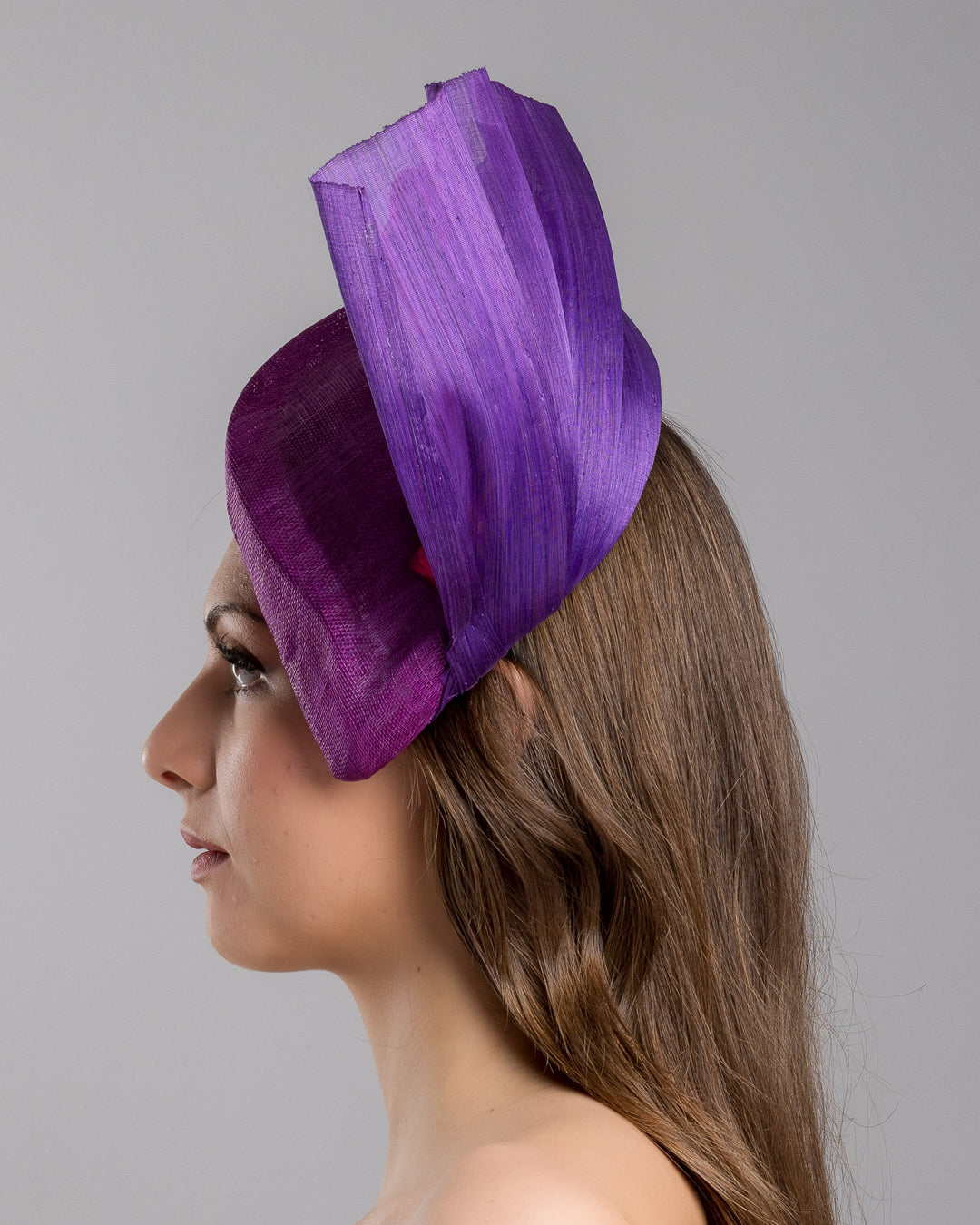 The model is showcasing the left side close up of the West is a purple side percher style fascinator curving to the left of the head framing the right side of the face. Decorated with a variety of pink and purple shaded cherry flowers and completed with a purple pleaded fan in the back. Designed and made by Merve Bayindir Millinery.
