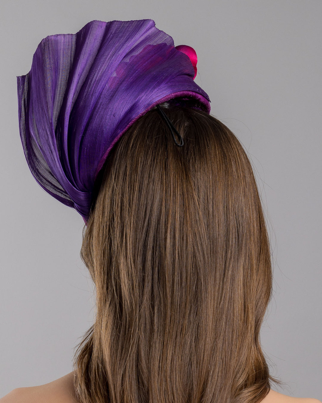 The model is showcasing the back side close up of the West is a purple side percher style fascinator curving to the left of the head framing the right side of the face. Decorated with a variety of pink and purple shaded cherry flowers and completed with a purple pleaded fan in the back. Designed and made by Merve Bayindir Millinery.
