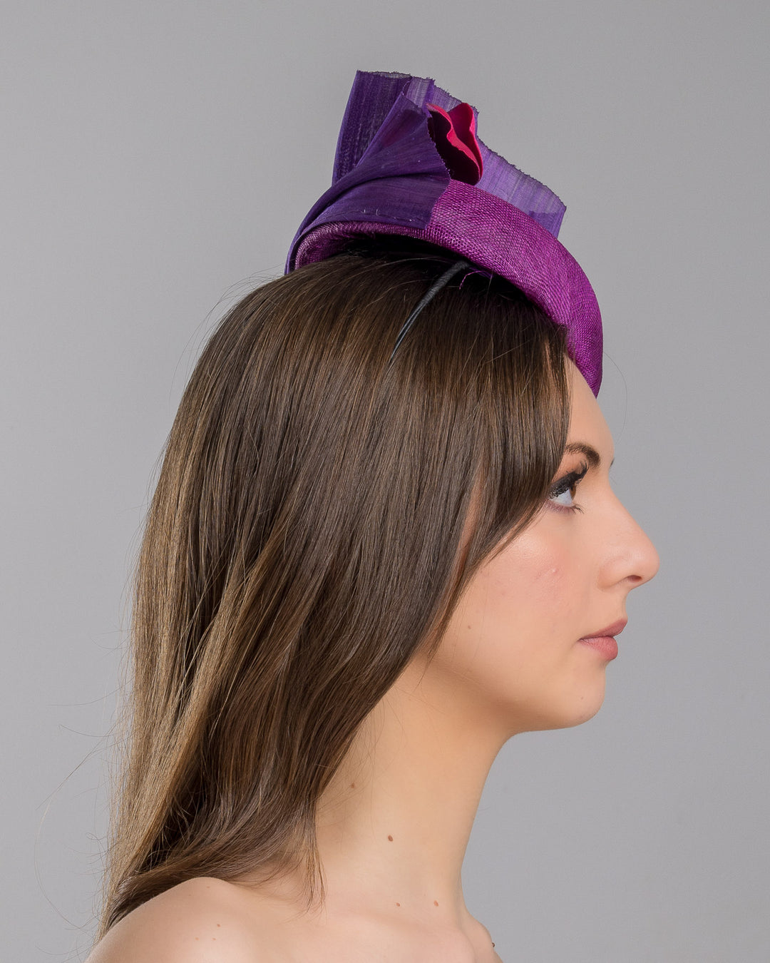 The model is showcasing the right side close up of the West is a purple side percher style fascinator curving to the left of the head framing the right side of the face. Decorated with a variety of pink and purple shaded cherry flowers and completed with a purple pleaded fan in the back. Designed and made by Merve Bayindir Millinery.
