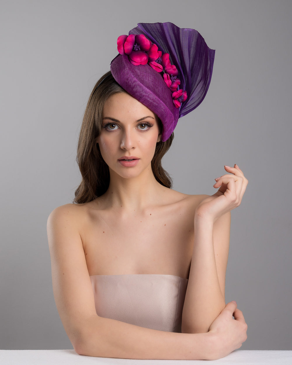 The female model is posing, wearing, West is a purple side percher style fascinator curving to the left of the head framing the right side of the face. Decorated with a variety of pink and purple shaded cherry flowers and completed with a purple pleaded fan in the back. Designed and made by Merve Bayindir Millinery.
