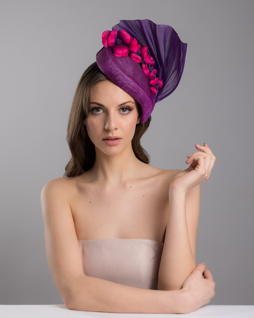 The female model is posing, wearing, West is a purple side percher style fascinator curving to the left of the head framing the right side of the face. Decorated with a variety of pink and purple shaded cherry flowers and completed with a purple pleaded fan in the back. Designed and made by Merve Bayindir Millinery.
