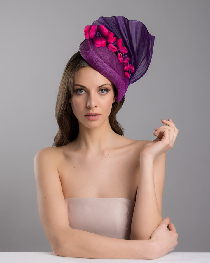 The female model is posing, wearing, West is a purple side percher style fascinator curving to the left of the head framing the right side of the face. Decorated with a variety of pink and purple shaded cherry flowers and completed with a purple pleaded fan in the back. Designed and made by Merve Bayindir Millinery.

