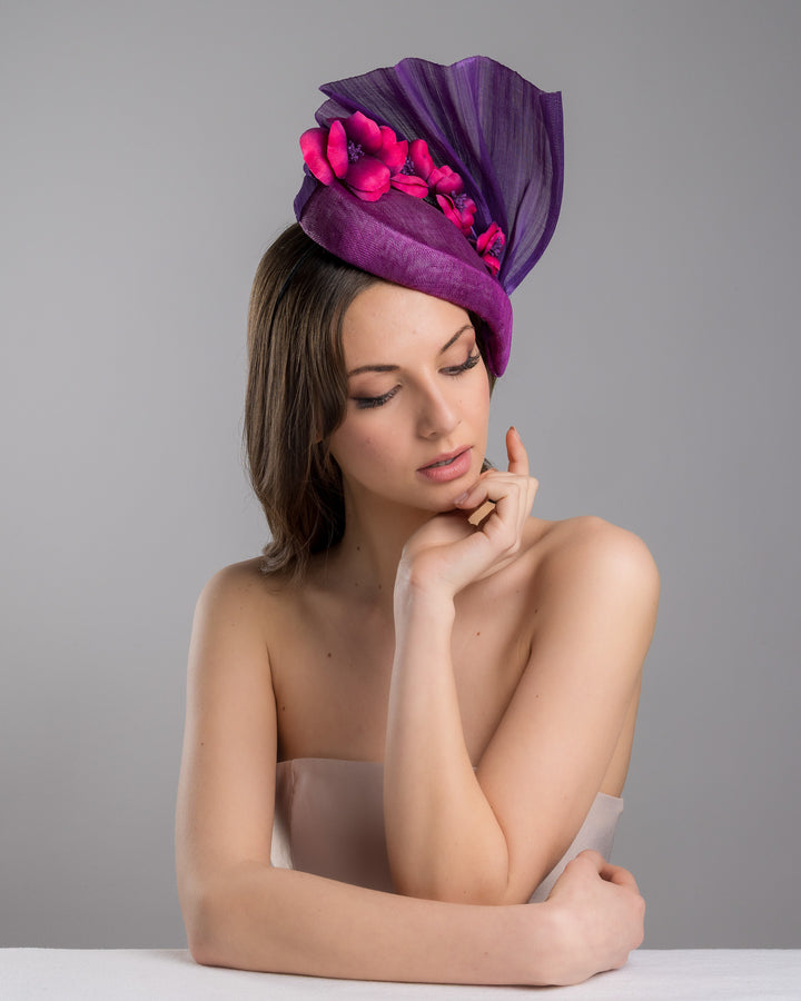 The female model is posing, wearing, West is a purple side percher style fascinator curving to the left of the head framing the right side of the face. Decorated with a variety of pink and purple shaded cherry flowers and completed with a purple pleaded fan in the back. Designed and made by Merve Bayindir Millinery.
