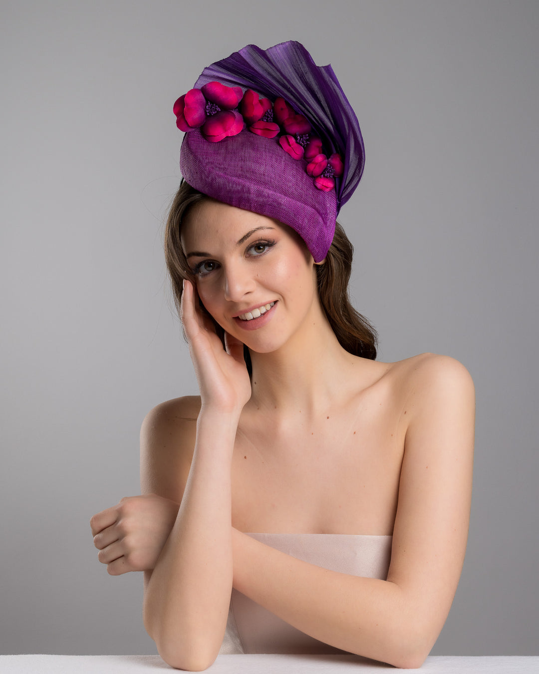 The female model is posing, wearing, West is a purple side percher style fascinator curving to the left of the head framing the right side of the face. Decorated with a variety of pink and purple shaded cherry flowers and completed with a purple pleaded fan in the back. Designed and made by Merve Bayindir Millinery.
