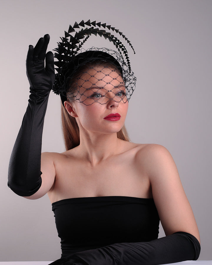 Model is showcasing the bottom front right angle of the black velvet high headband is decorated with widdow style veil, adorned with custom trimmed black rooster feathers curving on top of the headband. The end of the feathers is embellished with black crystal cluster where they meet with the headband.