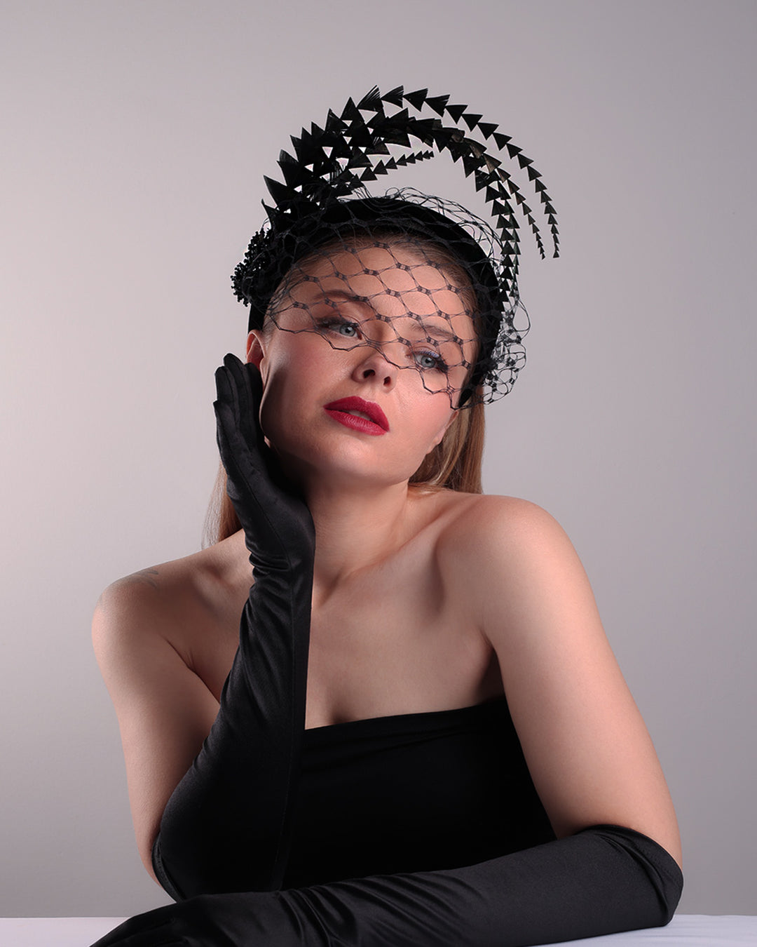 Model is showcasing the bottom front angle of the black velvet high headband is decorated with widdow style veil, adorned with custom trimmed black rooster feathers curving on top of the headband. The end of the feathers is embellished with black crystal cluster where they meet with the headband.