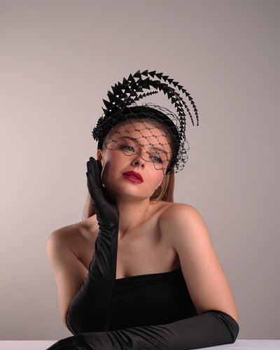 Model is showcasing the bottom front angle of the black velvet high headband is decorated with widdow style veil, adorned with custom trimmed black rooster feathers curving on top of the headband. The end of the feathers is embellished with black crystal cluster where they meet with the headband.