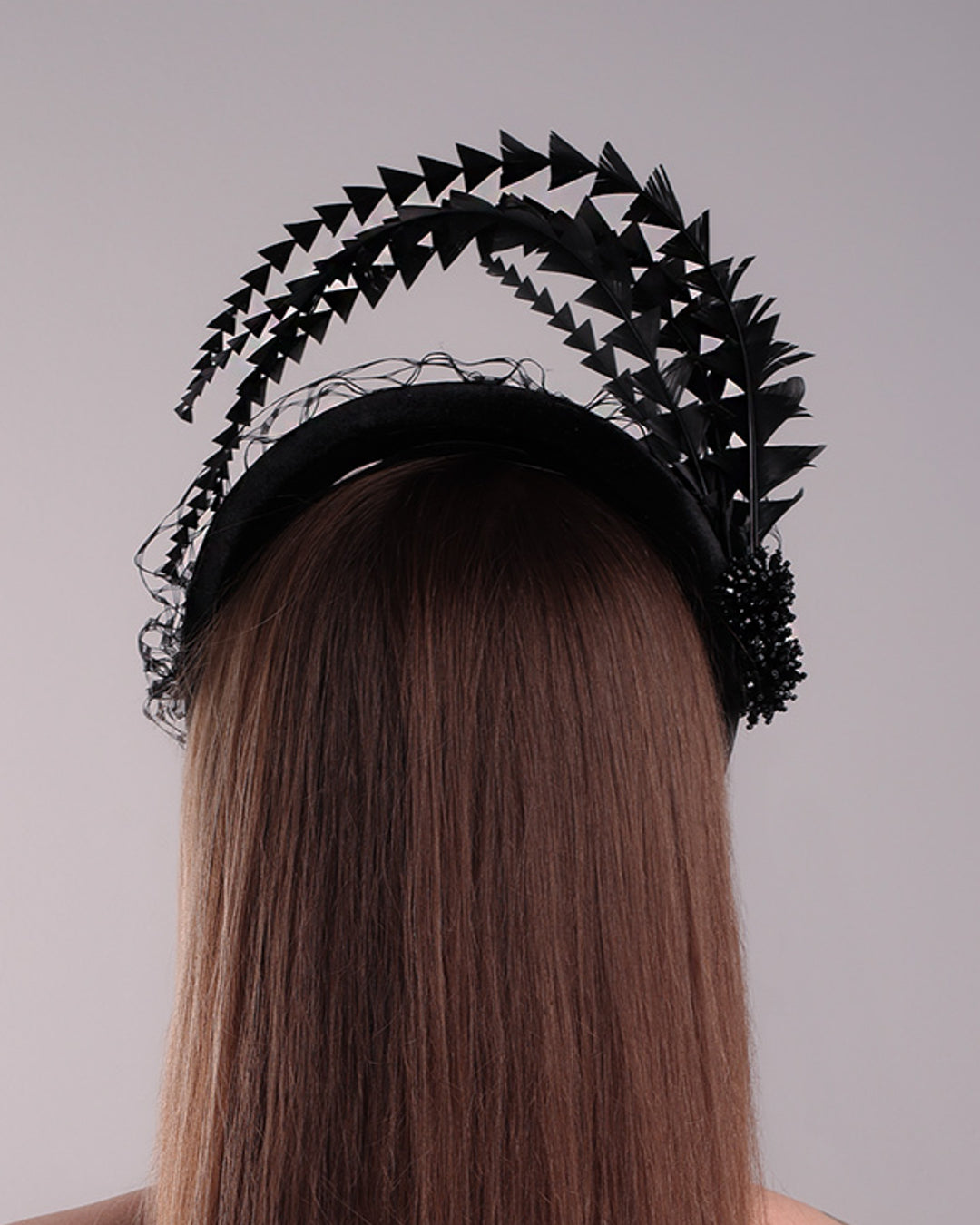 Model is showcasing the back side of the black velvet high headband is decorated with widdow style veil, adorned with custom trimmed black rooster feathers curving on top of the headband. The end of the feathers is embellished with black crystal cluster where they meet with the headband.