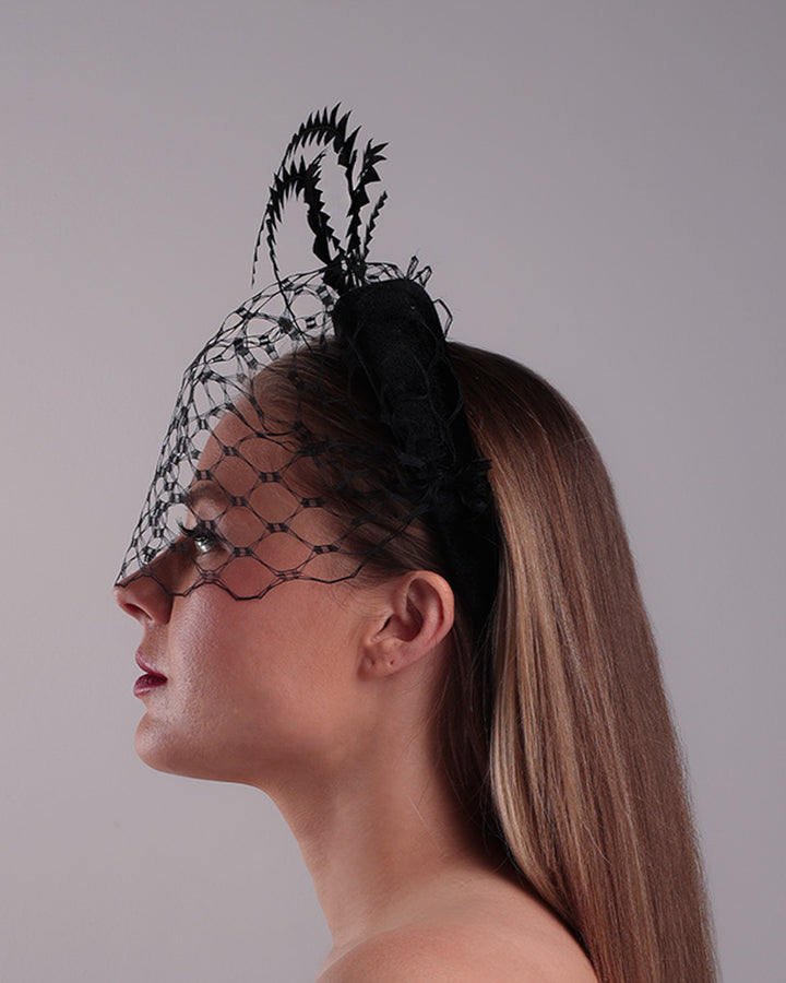 Model is showcasing the left side of the black velvet high headband is decorated with widdow style veil, adorned with custom trimmed black rooster feathers curving on top of the headband. The end of the feathers is embellished with black crystal cluster where they meet with the headband.