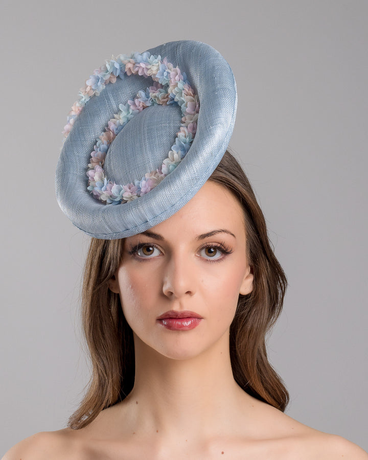 The model is showcasing the front side close up of the light blue, curved oval shaped high fascinator decorated with pastel colored organza flowers that circle the center and extend towards the back, providing additional flare. Designed and made by Merve Bayindir Millinery.
 
