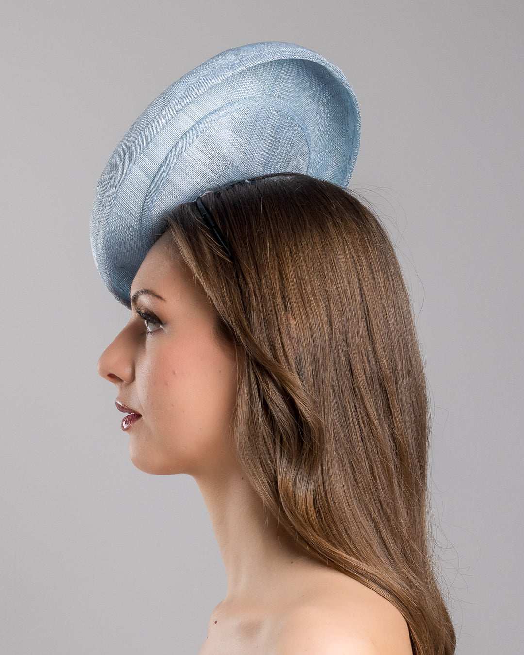The model is showcasing the left side close up of the light blue, curved oval shaped high fascinator decorated with pastel colored organza flowers that circle the center and extend towards the back, providing additional flare. Designed and made by Merve Bayindir Millinery.
 
