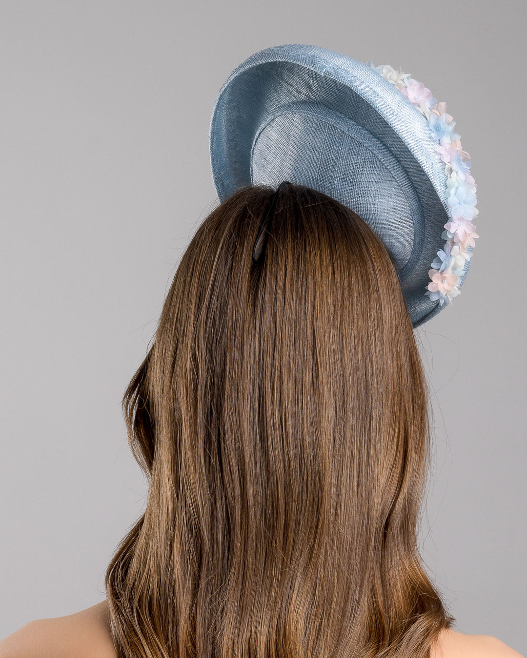 The model is showcasing the back side close up of the light blue, curved oval shaped high fascinator decorated with pastel colored organza flowers that circle the center and extend towards the back, providing additional flare. Designed and made by Merve Bayindir Millinery.
 
