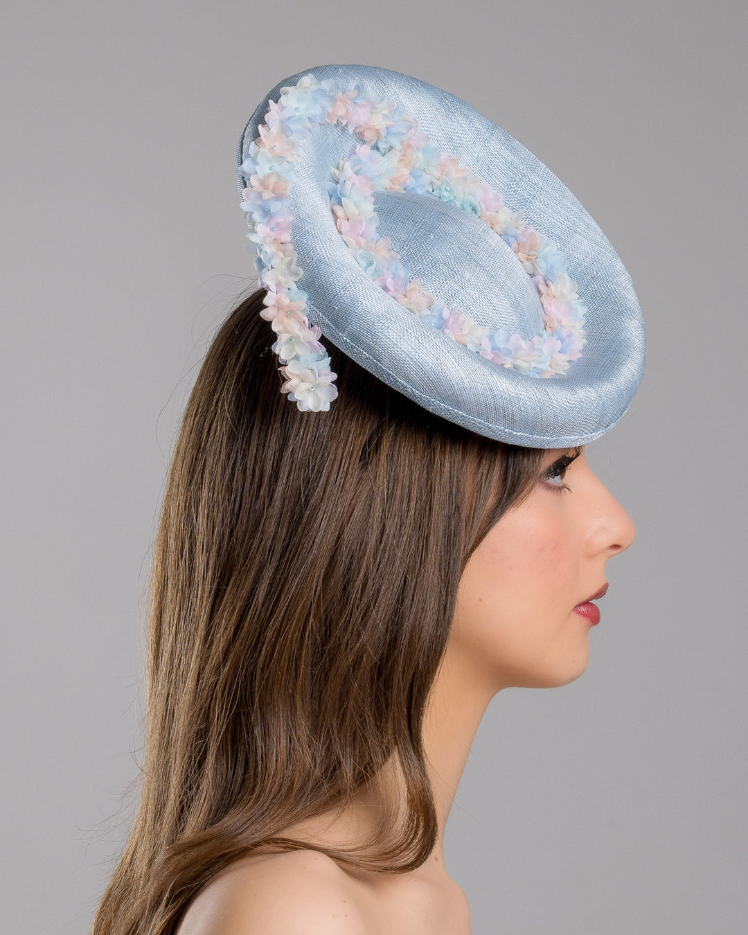 The model is showcasing the right side close up of the light blue, curved oval shaped high fascinator decorated with pastel colored organza flowers that circle the center and extend towards the back, providing additional flare. Designed and made by Merve Bayindir Millinery.
 
