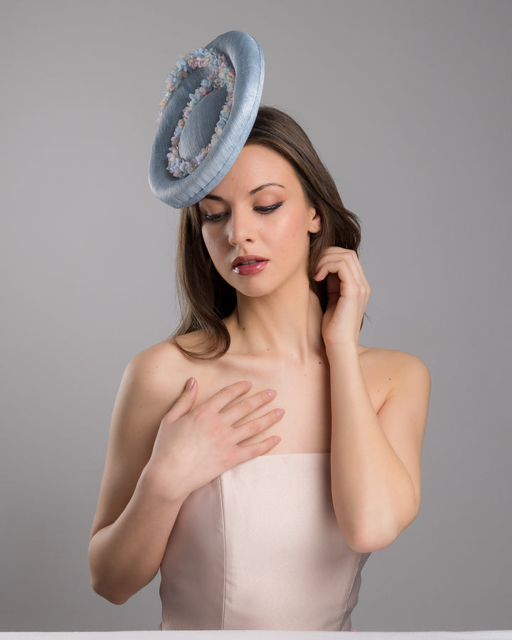 The female model is posing, wearing Wren,the light blue, curved oval shaped high fascinator decorated with pastel colored organza flowers that circle the center and extend towards the back, providing additional flare. Designed and made by Merve Bayindir Millinery.
 
