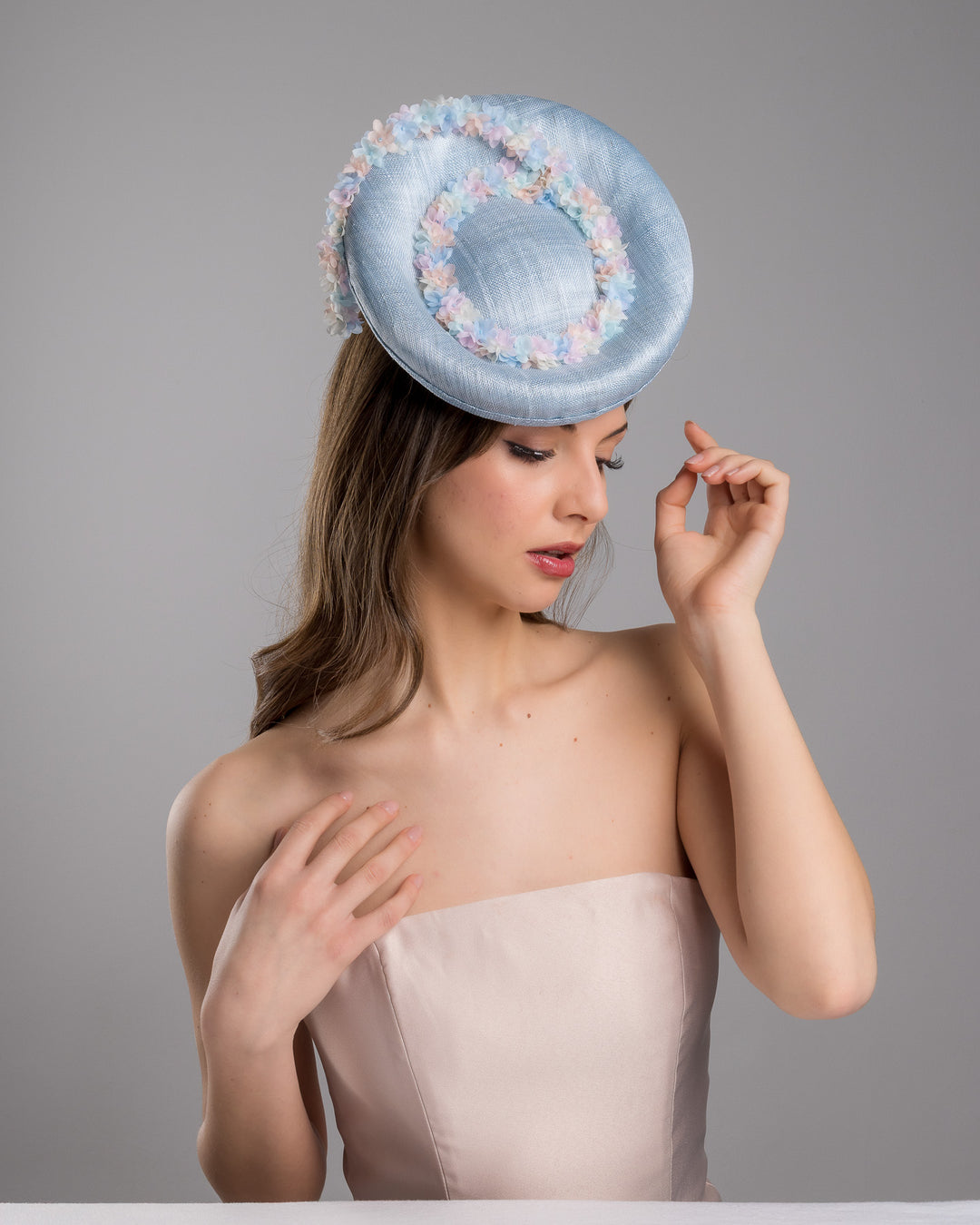 The female model is posing, wearing Wren,the light blue, curved oval shaped high fascinator decorated with pastel colored organza flowers that circle the center and extend towards the back, providing additional flare. Designed and made by Merve Bayindir Millinery.
 
