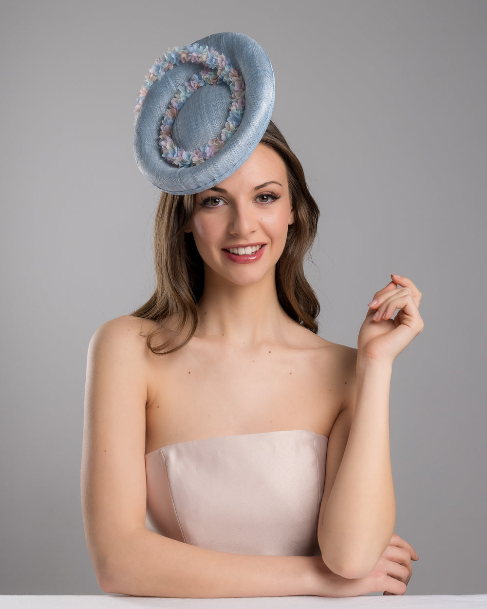 The female model is posing, wearing Wren,the light blue, curved oval shaped high fascinator decorated with pastel colored organza flowers that circle the center and extend towards the back, providing additional flare. Designed and made by Merve Bayindir Millinery.
 
