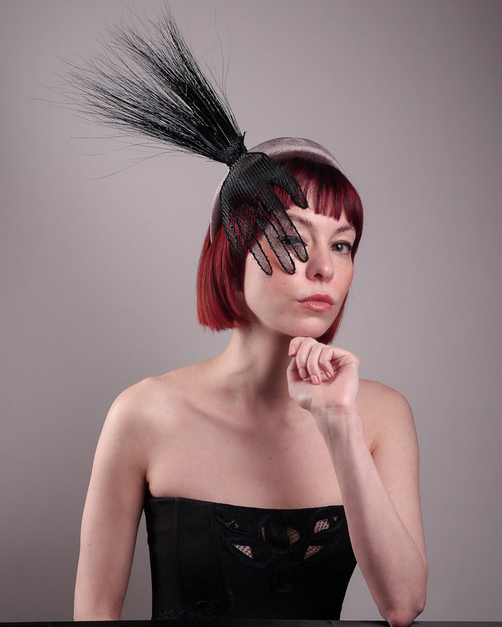 The model showcases the front right side of the Zephyr grey velvet headband with the hand-shaped wire black glove fitted with crinoline cascades dramatically across the face, balanced by a bursting spray of sleek horsehair. Designed by Merve Bayindir.