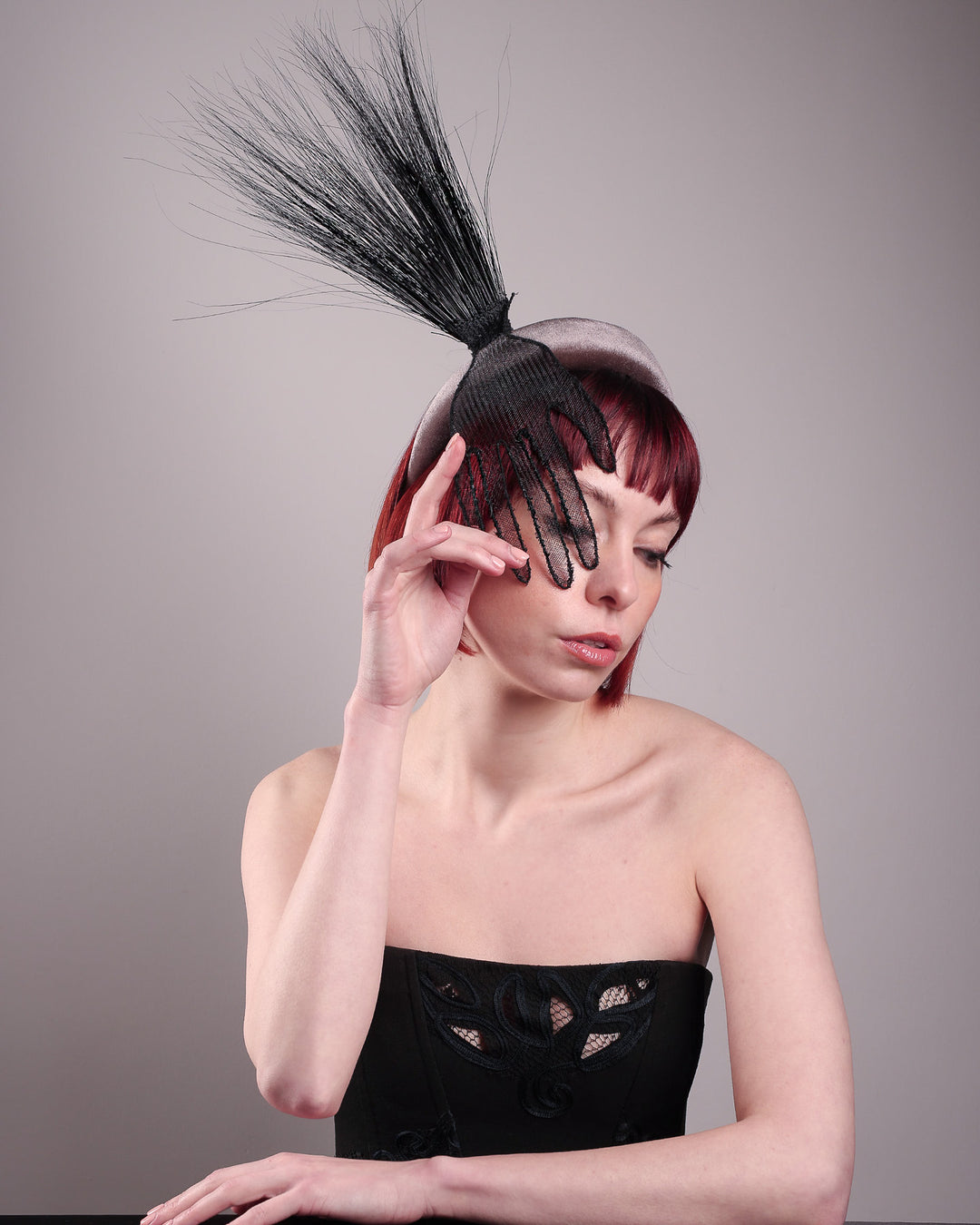 The model showcases the front right side from above of the Zephyr grey velvet headband with the hand-shaped wire black glove fitted with crinoline cascades dramatically across the face, balanced by a bursting spray of sleek horsehair. Designed by Merve Bayindir.