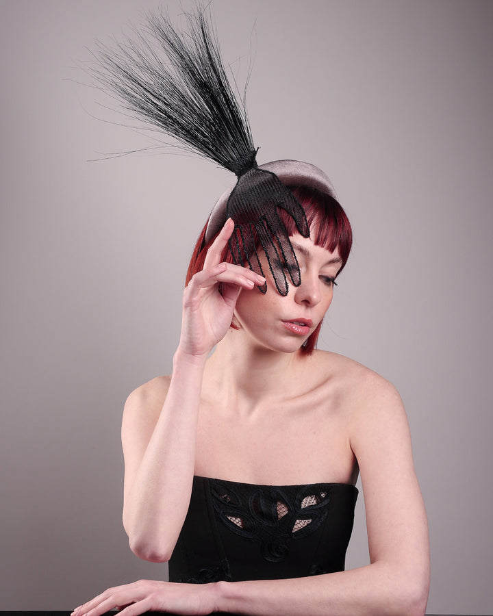 The model showcases the front right side from above of the Zephyr grey velvet headband with the hand-shaped wire black glove fitted with crinoline cascades dramatically across the face, balanced by a bursting spray of sleek horsehair. Designed by Merve Bayindir.