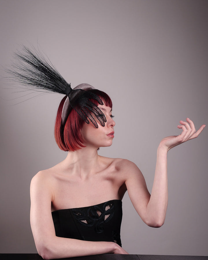 The model showcases the right side of the Zephyr grey velvet headband with the hand-shaped wire black glove fitted with crinoline cascades dramatically across the face, balanced by a bursting spray of sleek horsehair. Designed by Merve Bayindir.