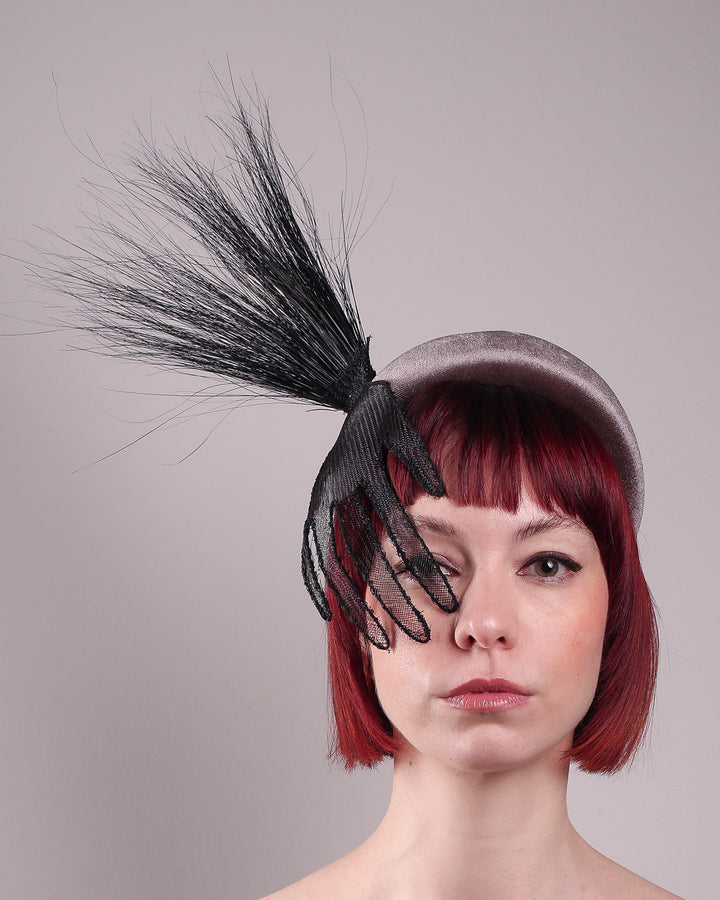 The model showcases the front close up of the Zephyr grey velvet headband with the hand-shaped wire black glove fitted with crinoline cascades dramatically across the face, balanced by a bursting spray of sleek horsehair. Designed by Merve Bayindir.