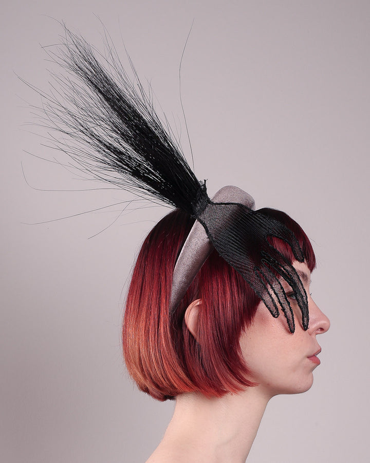 The model showcases the right side close up of the Zephyr grey velvet headband with the hand-shaped wire black glove fitted with crinoline cascades dramatically across the face, balanced by a bursting spray of sleek horsehair. Designed by Merve Bayindir.