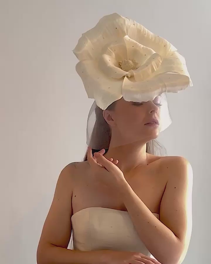 Model is showcasing a large white hand made silk abaca flower, decorated with silver crystals, placed one a button shaped white sinamay fascinator from the left side A white crinoline veil is placed between the hat and the flower to cover the face. The button base is fixed on a custom headband and secured with an additional elastic.