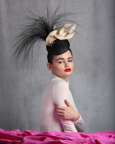 The female model is wearing a show-stopping black button-shaped high fascinator. The fascinator is decorated with multiple ivory swirling ribbon-looking jinns, giving the impression of flying ribbons. One side of the swirls attached to the fascinator is embellished with mother of pearls, and the back of the fascinator has long black burnt peacock feathers.