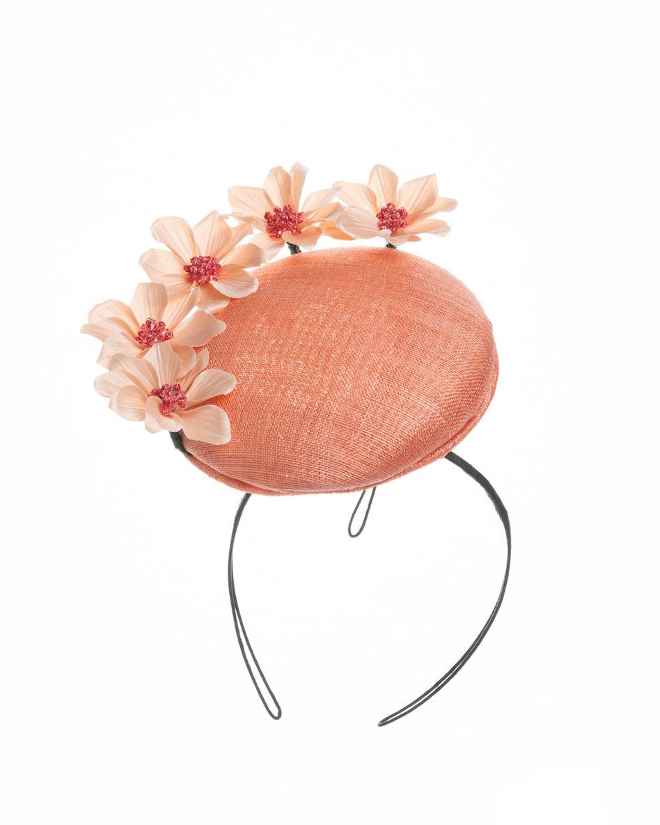 A coral coloured tall button shaped fascinator attached towards the right side of a headband. The fascinator is decorated from right side to the top with lighter coral coloured five flowers. Designed and made by Merve Bayindir millinery.