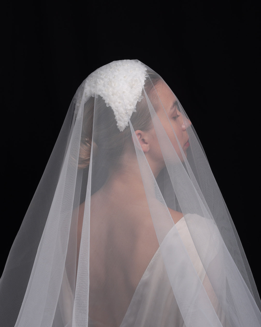  The model showcases the back of a custom-shaped bridal headpiece embellished with small organza flowers and pearl beads, covered with a sheer long veil.